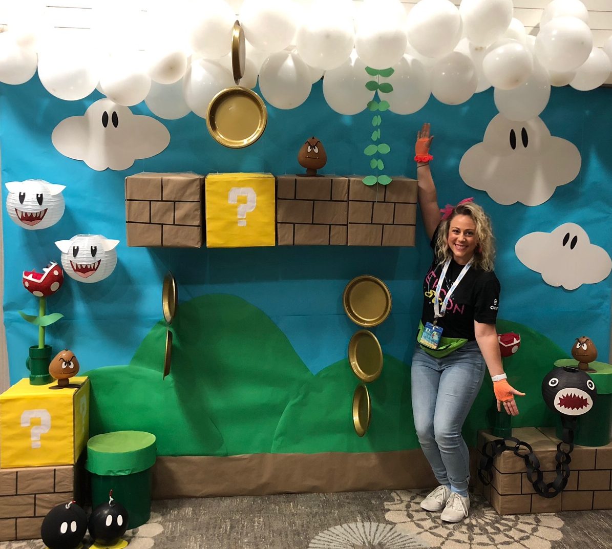 Jen standing in front of a handmade Mario-inspired photo op scene at DevCon 2019, with white balloon clouds and yellow question mark boxes